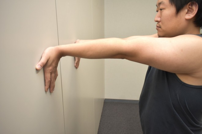 1. 壁の前に立つ 2. 手首を裏返した状態(指が下になる)で壁に手のひらをつける 3. 頭を壁に近づけて前重心になる
